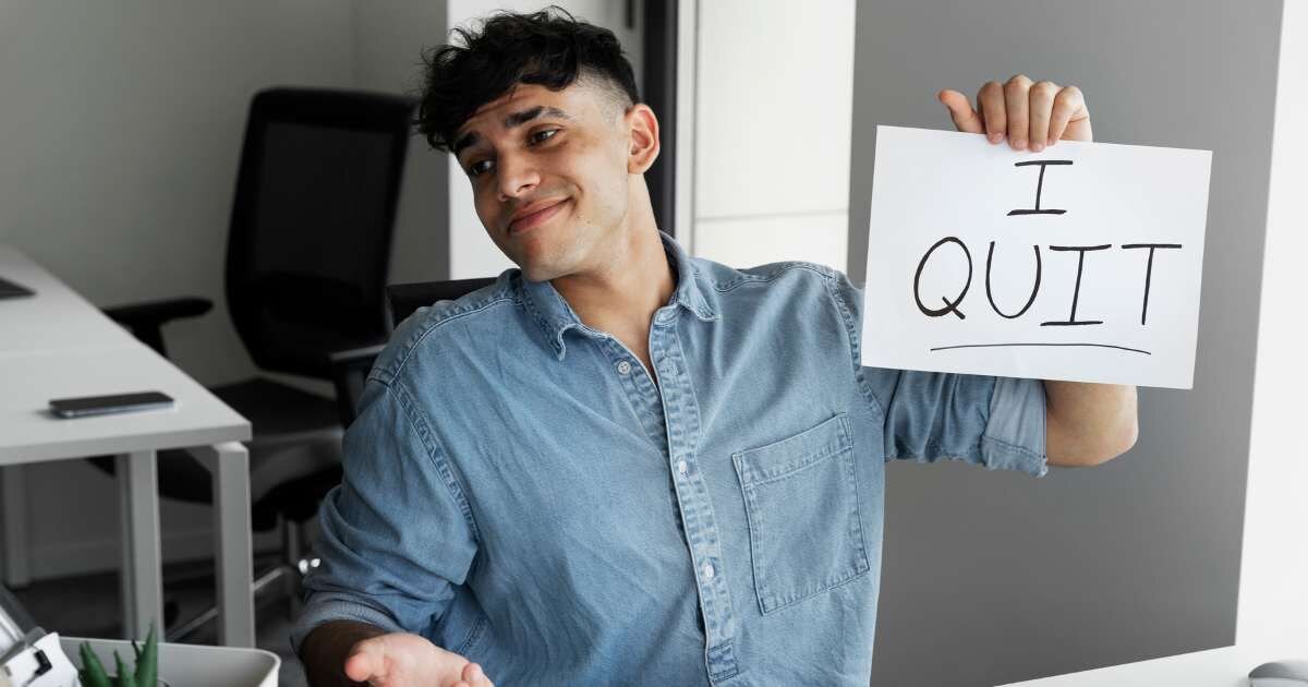 young man holding up an "I quit" sign while sitting at an office desk 